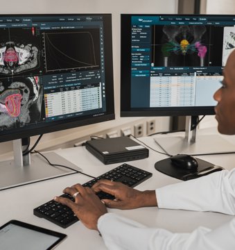 A radiologist sitting at a desk, looking at two computer screens showing scan software
